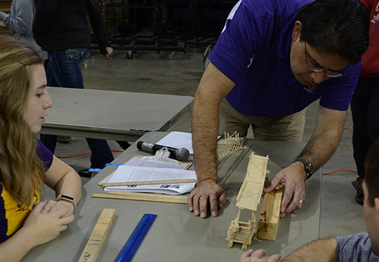 Link to Learn more; Photo of competition judges inspecting a team's toothpick bridge before testing.