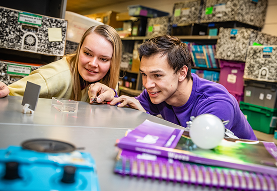 Link, Two future educators are using materials from a kit about lasers and light waves.  They are surrounded by shelves of science kits and supplemental curriculum.